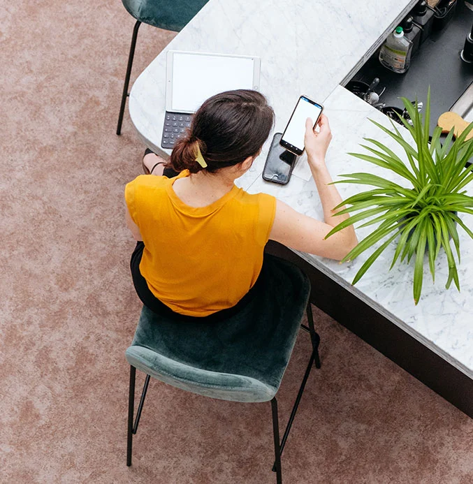 A woman sits at a marble kitchen island, holding a cell phone and working on her laptop.